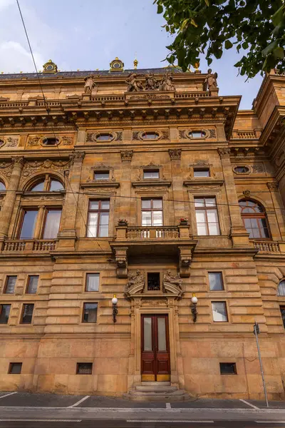 Detaylı oymaları, birden fazla penceresi ve belirgin girişi olan, parçalı bulutlu bir gökyüzüne karşı duran büyük, tarihi bir bina..