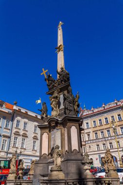 Altın haçla süslenmiş ve heykellerle çevrili uzun, süslü bir sütun açık mavi gökyüzüne karşı belirgin bir şehir meydanında duruyor, arka planda binalar görünüyor..