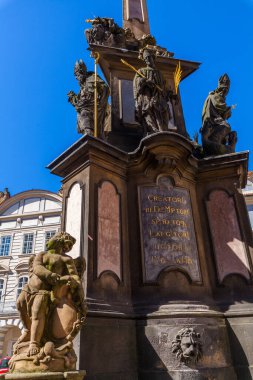 Karmaşık heykeller ve yazıtlarla süslenmiş, açık mavi gökyüzü ve bulanık binaların zeminine kurulmuş tarihi bir veba sütununun yakın çekimi..