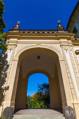 Üzerinde dekoratif heykeller olan kemerli bir kapının alçak açılı çekimi, parlak mavi gökyüzüne karşı ağaçların ve binaların manzarasını çerçeveliyor..
