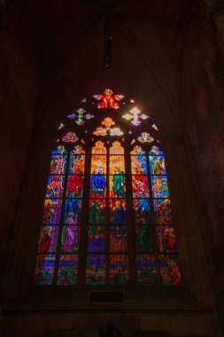 Karmaşık tasarımları ve figürleri olan canlı bir vitray. Güneş ışığıyla aydınlatılmış. Gotik bir kemer yolunun karanlık iç kısımlarında parlıyor..