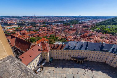 Geniş bir Avrupa şehrine ev sahipliği yapan yüksek açılı bir şehir, kırmızı kiremitli çatıları olan, yüksek bir noktadan görünen, ön planda bir taş balyoz, açık mavi bir gökyüzü altında.