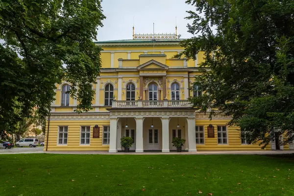 Sütunlu girişi ve dekoratif detayları olan soluk sarı cepheli, büyük, simetrik bir bina, bulutlu bir gökyüzünün altındaki yeşil bir parkta belirgin bir şekilde duruyor..