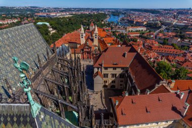 Karmaşık mimari detaylar ve kırmızı çatılı binaların yer aldığı yüksek açılı bir çatı manzarası, nehir ve mavi gökyüzünün altındaki köprülere bakan Avrupa şehir manzarası.