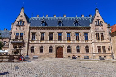 Süslü cepheli tarihi bir bina, birden fazla pencere, öne çıkan bir giriş, ve önünde parlak mavi bir gökyüzünün altında bir kaldırım taşı kare..