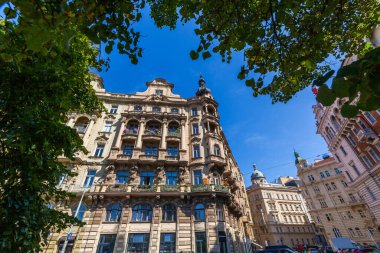 Dekoratif balkonlar, heykeller ve açık mavi gökyüzünün altında kısmen yeşil yapraklarla çerçevelenmiş pencere detaylarıyla süslü bir binanın ayrıntılı bir görüntüsü..