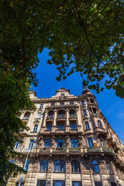 Dekoratif balkonlar, heykeller ve açık mavi gökyüzünün altında kısmen yeşil yapraklarla çerçevelenmiş pencere detaylarıyla süslü bir binanın ayrıntılı bir görüntüsü..