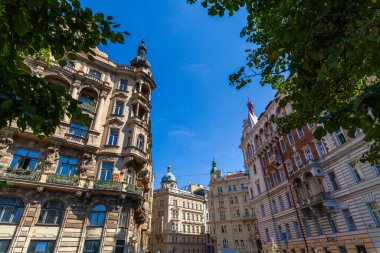 Açık mavi gökyüzüne karşı farklı mimari tarzlara sahip, ön plandaki yeşil yapraklarla çerçevelenmiş canlı bir şehir manzarası..