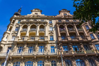 Dekoratif balkonlar, heykeller ve açık mavi gökyüzünün altında kısmen yeşil yapraklarla çerçevelenmiş pencere detaylarıyla süslü bir binanın ayrıntılı bir görüntüsü..