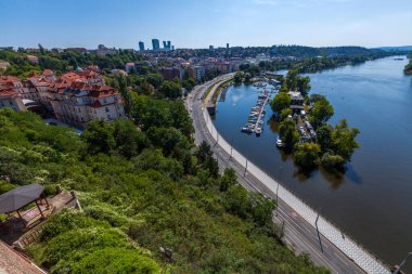 Geniş bir nehrin yüksek açılı görüntüsü kırmızı çatılı binalar, köprüler ve açık mavi gökyüzünün altındaki yeşil alanlar üzerinden akıyor..
