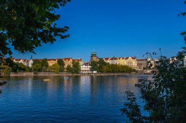 Mavi gökyüzünü yansıtan geniş bir nehir, kıyıları kaplayan renkli binalar, ön planda ağaçlar ve suyu noktalayan küçük tekneler..