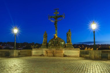 Alacakaranlık 'ta, arka planda derin mavi bir gökyüzünün altında, sokak lambalarıyla aydınlatılmış, haç ve heykellerle süslenmiş merkezi bir dini anıtın yer aldığı taş bir köprü.