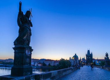 Bir haç tutan bir adam heykeli, alacakaranlık gökyüzüne karşı siluet, kırmızı çatılı binalara bakan bir şehir manzarası ve uzakta büyük bir bina kompleksi, Charles Köprüsü.