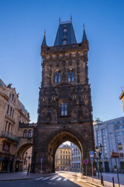 Karanlık kulesi ve kemerli geçidi olan uzun, gotik bir kule, açık mavi gökyüzünün altında binaları ve tabelaları olan bir şehir meydanında belirgin bir şekilde duruyor..