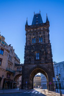 Karanlık kulesi ve kemerli geçidi olan uzun, gotik bir kule, açık mavi gökyüzünün altında binaları ve tabelaları olan bir şehir meydanında belirgin bir şekilde duruyor..