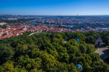 Yüksek görüş açısına sahip panoramik bir şehir manzarası, kırmızı çatılı binalar, büyük bir yapı kompleksi ve açık mavi gökyüzünün altında uzak bir ufuk çizgisi olan geniş bir şehri gösteriyor..