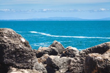 Huzurlu bir okyanus manzarası. Parıldayan mavi su ufka doğru uzanıyor. Önplanda kayalık oluşumlar ve dalgalardan yükselen yalnız bir kaya.