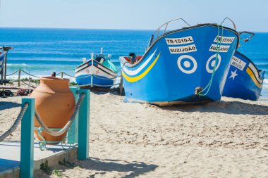 This image captures a serene coastal scene with brightly painted fishing boats resting on a sandy beach. A terracotta-colored clay pot and rope fencing in the foreground add a rustic touch to the scene, reinforcing the fishing village vibe. The entir clipart