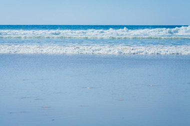 Bu görüntü sakin bir okyanus sahnesini yakalar ve dalgalar kıyıya doğru ilerler. Ufuk, mavi okyanusun daha açık gökyüzüne karışmasıyla görüntünün üstüne doğru uzanıyor. Ön plandaki su pürüzsüz, yumuşak ışığı yansıtıyor.