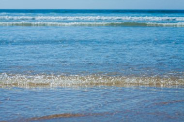 Bu görüntü sakin bir okyanus sahnesini yakalar ve dalgalar kıyıya doğru ilerler. Ufuk, mavi okyanusun daha açık gökyüzüne karışmasıyla görüntünün üstüne doğru uzanıyor. Ön plandaki su pürüzsüz, yumuşak ışığı yansıtıyor.