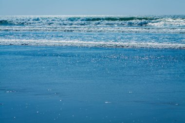 Bu görüntü sakin bir okyanus sahnesini yakalar ve dalgalar kıyıya doğru ilerler. Ufuk, mavi okyanusun daha açık gökyüzüne karışmasıyla görüntünün üstüne doğru uzanıyor. Ön plandaki su pürüzsüz, yumuşak ışığı yansıtıyor.