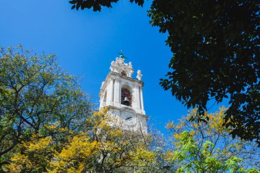 Uzun, zarif bir beyaz saat kulesi. Karmaşık mimari detayları var. Muhtemelen bir kilise ya da tarihi binanın parçası. Kuledeki çan görünür, dekoratif taş işçiliğiyle çerçevelenmiş ve süslü süslemeler ve heykellerle süslenmiş. Saat yüzü.