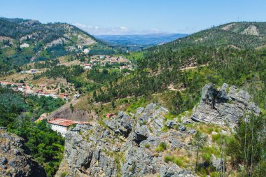 Bereketli yeşillikler, kayalık uçurumlar ve uzak bir köprü ile çevrili dağlık bir arazide yuva yapmış küçük bir köyün panoramik görüntüsü doğanın ve kırsal yaşamın barışçıl güzelliğini yakalıyor.