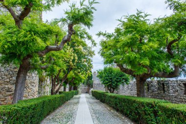 Manikürlü çitlerle ve yemyeşil ağaçlarla kaplı, antik taş duvarlara ve kulelere uzanan manzaralı taş bir yol parçalı bulutlu gökyüzünün altında huzurlu ve tarihi bir atmosfer sunuyor.
