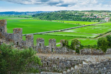 Sarmaşıklarla kaplı eski bir taş duvar geniş yeşil alanları ve yakınlardaki bir köyü gözden geçirir, tarihi yapıyı bulutlu bir gökyüzünün altındaki canlı kırsal manzarayla harmanlar.