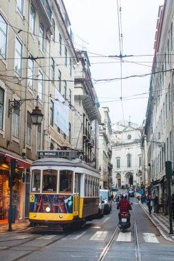 Lizbon 'un Baixa bölgesinin klasik bir sahnesi, ikonik sarı tramvaylarından biri dar, kaldırımlı caddeler boyunca seyahat ediyor. Tramvay kırmızı ekose ceketli bir motosikletli tarafından yakından takip edilir. Her iki taraftaki binalar da uzun, tarihsel.