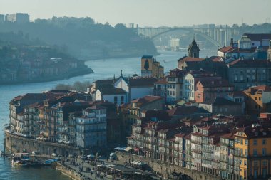 Renkli tarihi binaların rıhtıma doğru çağlayarak indiği manzaralı bir nehir manzarası, sisli tepelerden oluşan bir zemin ve nehir boyunca canlı kentsel yaşamın özünü yansıtan uzak bir köprü.