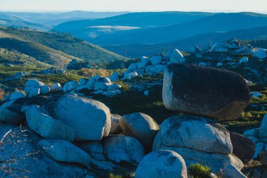 Gün batımında sakin bir dağ manzarası, geniş yuvarlak kayalar araziye saçılmış, yeşillik üzerine uzun gölgeler ve arka planda yuvarlanan tepeler