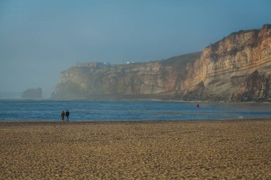 Bir çift, arka planda yüksek uçurumlar olan sakin kumlu bir sahil boyunca yürürken, sisli bir ufuktan gelen hafif dalgalar, sakin ve atmosferik bir kıyı manzarası yaratıyor.