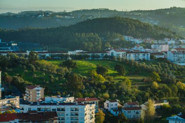 Ağaçlarla benekli ve konut binalarının yanında yuva yapmış, yuvarlak tepeli yemyeşil bir manzara. Arka planda, ormanlık tepeler yükselir ve önplandaki kentsel yapılara doğal bir tezat oluşturur..