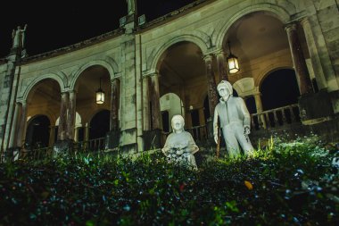 İki figürlü aydınlık heykeller gece yeşillikler arasında duruyor, kemerli bir sütunla karşı karşıya.