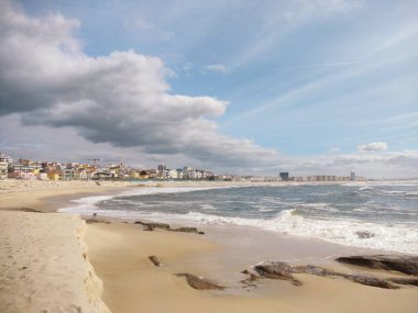 Parçalı bulutlu gökyüzünün altında hafif dalgalarla dolu kumlu bir sahil. Renkli alçak binalarla çevrili bir kasaba.