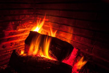 Şöminede yanan bir ateşin yakın çekim görüntüsü. Alevler odunların etrafında dans ediyor. Yoğun turuncu bir parıltı ve koyu kırmızı közler yayıyor. Sıcak, rahat bir atmosfer yaratıyor..