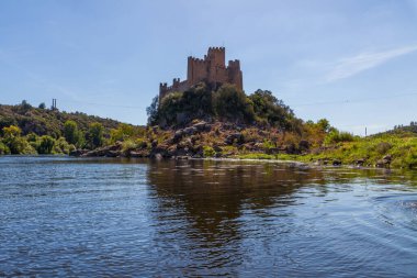 Tarihi bir şato, sakin nehir sularıyla çevrili kayalık bir adanın tepesinde berrak, parlak bir gökyüzünün altında yemyeşil tepeler arasında görkemli bir şekilde duruyor.
