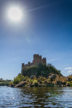 Tarihi bir şato, sakin nehir sularıyla çevrili kayalık bir adanın tepesinde berrak, parlak bir gökyüzünün altında yemyeşil tepeler arasında görkemli bir şekilde duruyor.