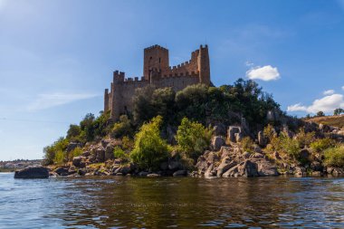 Tarihi bir şato, sakin nehir sularıyla çevrili kayalık bir adanın tepesinde berrak, parlak bir gökyüzünün altında yemyeşil tepeler arasında görkemli bir şekilde duruyor.