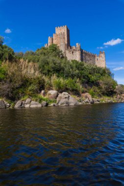 Tarihi bir şato, sakin nehir sularıyla çevrili kayalık bir adanın tepesinde berrak, parlak bir gökyüzünün altında yemyeşil tepeler arasında görkemli bir şekilde duruyor.
