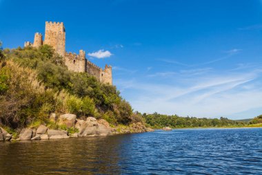 Tarihi bir şato, sakin nehir sularıyla çevrili kayalık bir adanın tepesinde berrak, parlak bir gökyüzünün altında yemyeşil tepeler arasında görkemli bir şekilde duruyor.