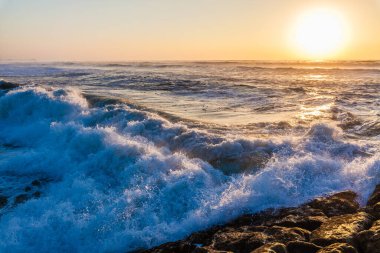 Güneş batarken okyanus dalgalarının gücü ve güzelliği. Ön planda büyük, köpüklü dalgalar kıyıya çarpıyor, her dalga doku ve enerjiyle zenginleşiyor..