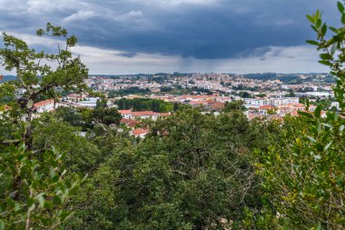 Kırmızı kiremitli çatıları olan, dramatik bulutlu gökyüzü altında yeşilliklerle çevrili büyüleyici bir kasabanın panoramik görüntüsü, kentsel yaşam ve doğal güzellik arasındaki zıtlığı yakalıyor.