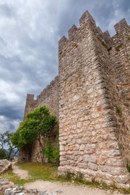 Çimenli bir yamaca tünemiş tarihi taş kale, dramatik bulutlu gökyüzünün altında yeşilliklerle çevrili, zamansız mimari güzelliğini vurguluyor.