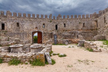 Çimenli bir yamaca tünemiş tarihi taş kale, dramatik bulutlu gökyüzünün altında yeşilliklerle çevrili, zamansız mimari güzelliğini vurguluyor.