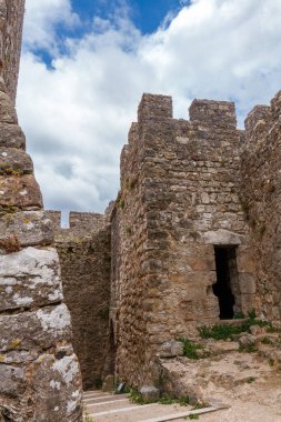 Çimenli bir yamaca tünemiş tarihi taş kale, dramatik bulutlu gökyüzünün altında yeşilliklerle çevrili, zamansız mimari güzelliğini vurguluyor.