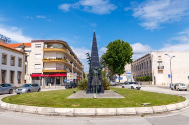 Yoğun bir kasabanın merkezinde canlı bir döner kavşak, yukarı doğru uzanan bronz figürlü modern dikilitaş şekilli bir heykel var.
