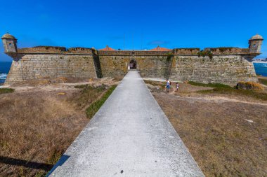 Yıpranmış taş duvarları ve girişe giden uzun bir yolu olan tarihi bir sahil kalesi, açık mavi gökyüzüne karşı kurulmuş.