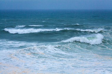 Yuvarlanan dalgalar ve beyaz köpüklerle dolu engin, güçlü bir okyanus yumuşak, sisli bir mavi gökyüzünün altında uzanır ve dinamik bir deniz manzarası yaratır.
